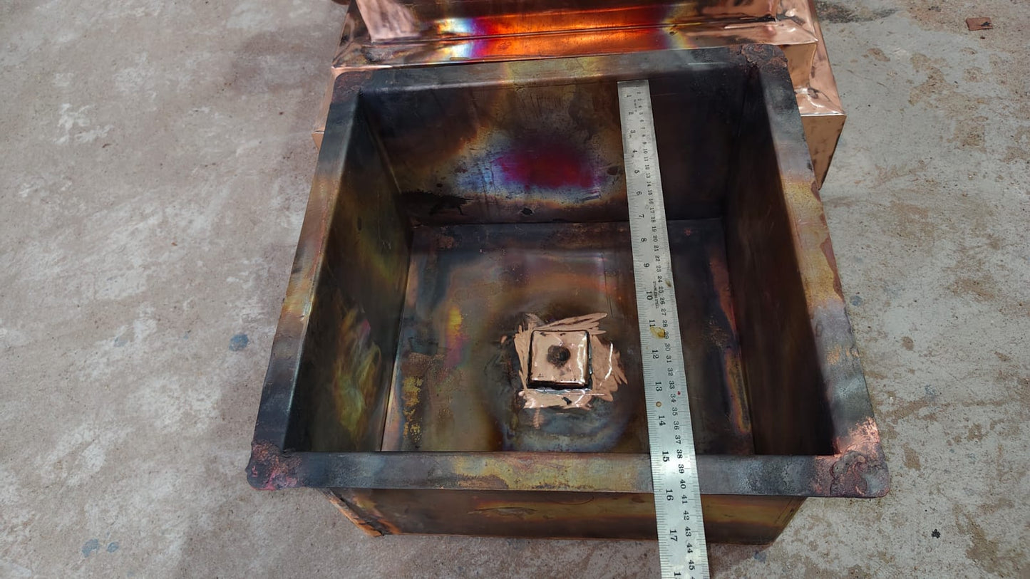A square metal container or furnace lid with a welded interior, showing rusted and oxidized surfaces, with a metal ruler placed inside for scale on a concrete floor.