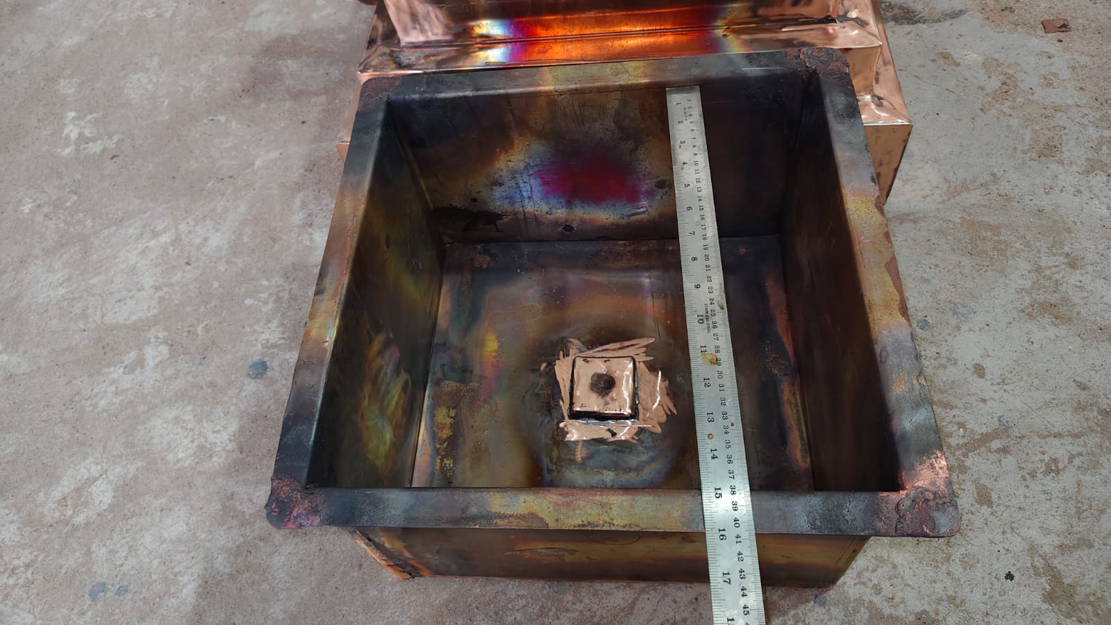A square metal container or furnace lid with a welded interior, showing rusted and oxidized surfaces, with a metal ruler placed inside for scale on a concrete floor.
